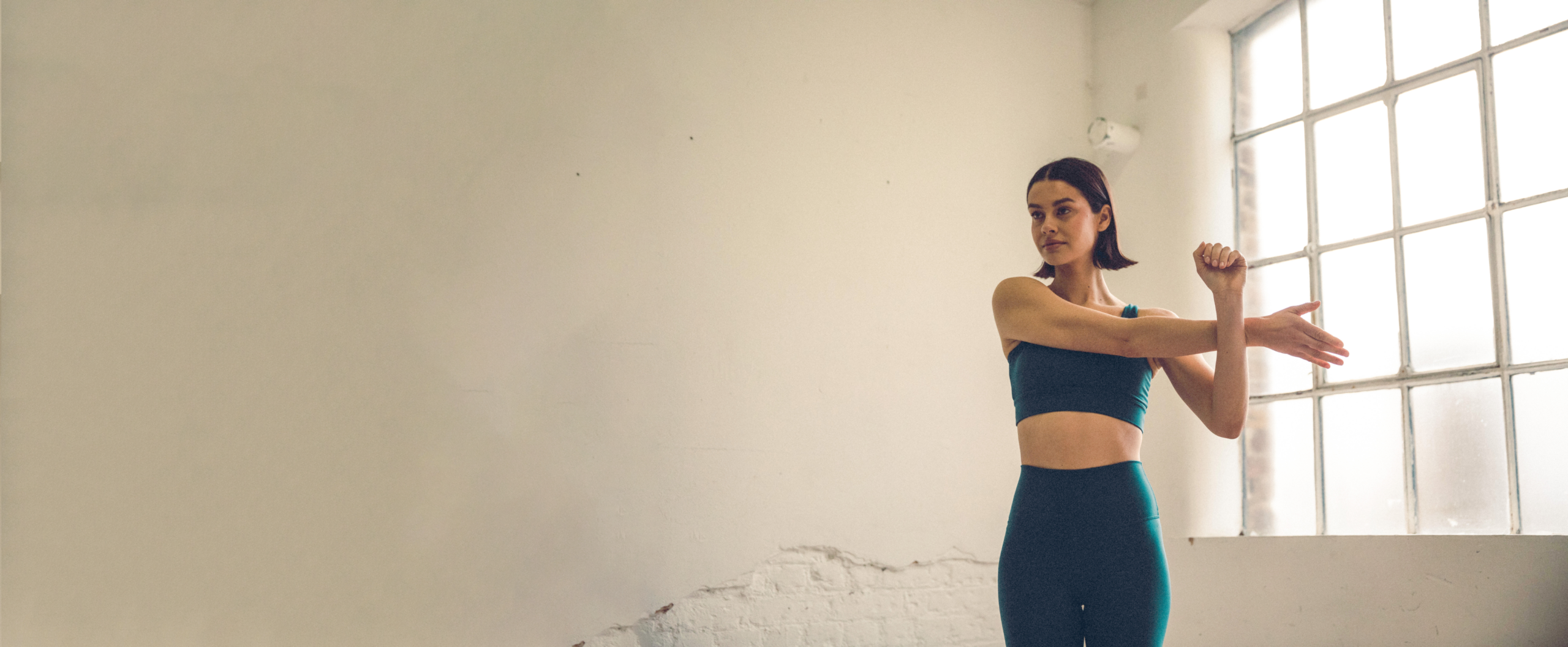Woman stretching in a Barrecore studio before class, wearing teal activewear in a minimalist, light-filled space.