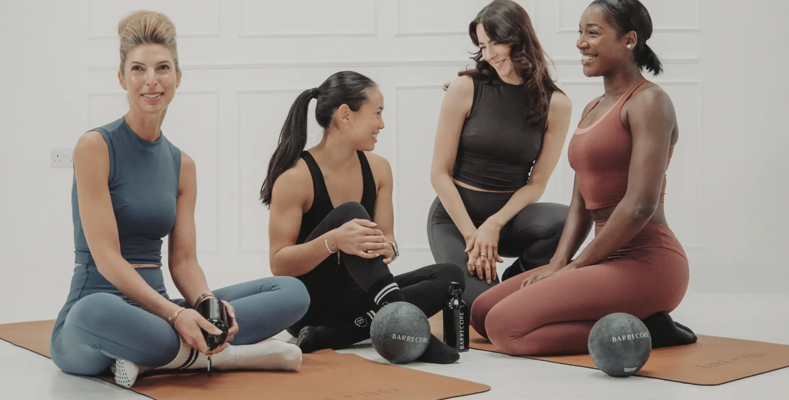 Four women sitting on mats in a Barrecore studio, chatting and smiling with Barrecore balls and water bottles beside them