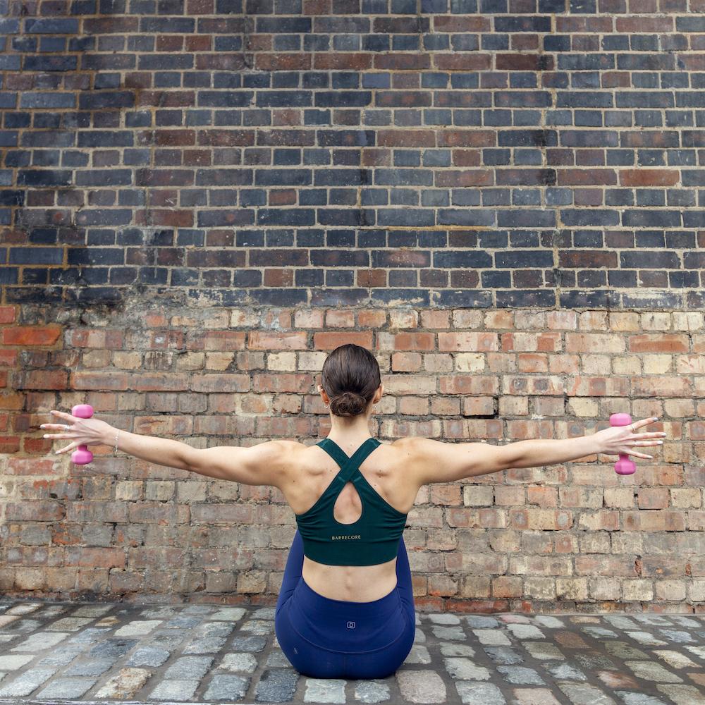 A women sits with her back to the camera sat on the floor in a barre position holding a small dumbbell in each hand