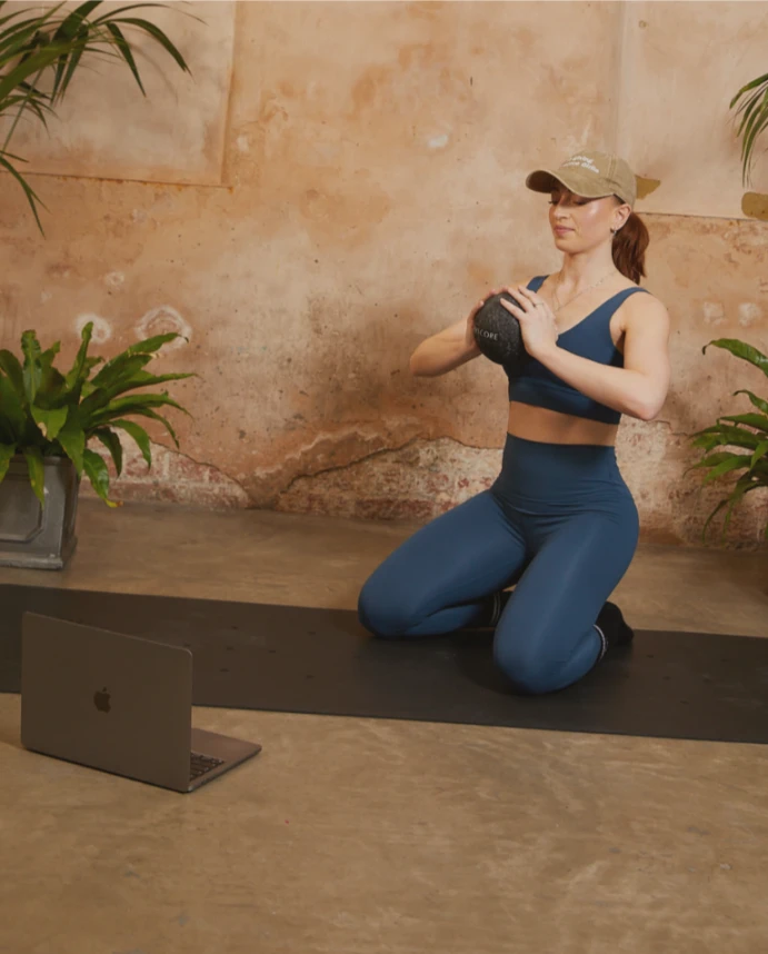 Woman in blue activewear following a virtual Barrecore class at home, kneeling with a weighted ball in front of a laptop.