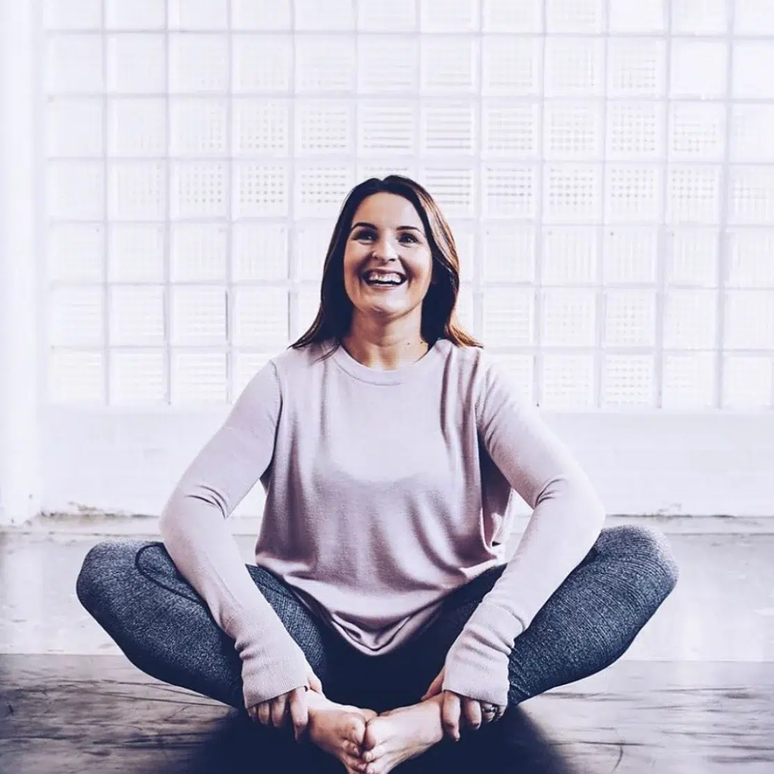 Sarah Emblow - a female with shoulder length dark hear wearing relaxed light leisurewear sat on the floor cross legged