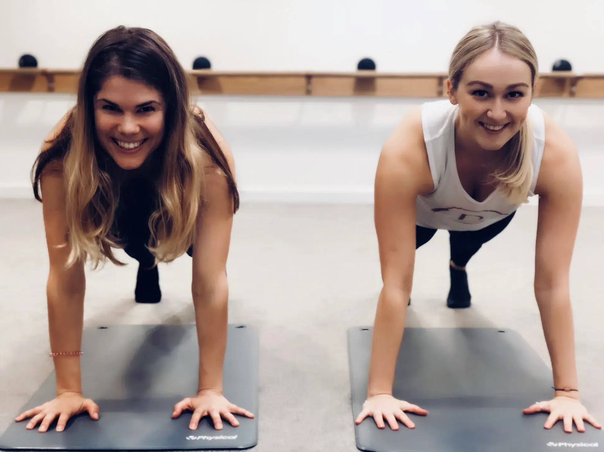 Two females, Chloe Eastwood and Madeleine Shaw are in a barre studio holding plank positions on exercise mats next to each other.