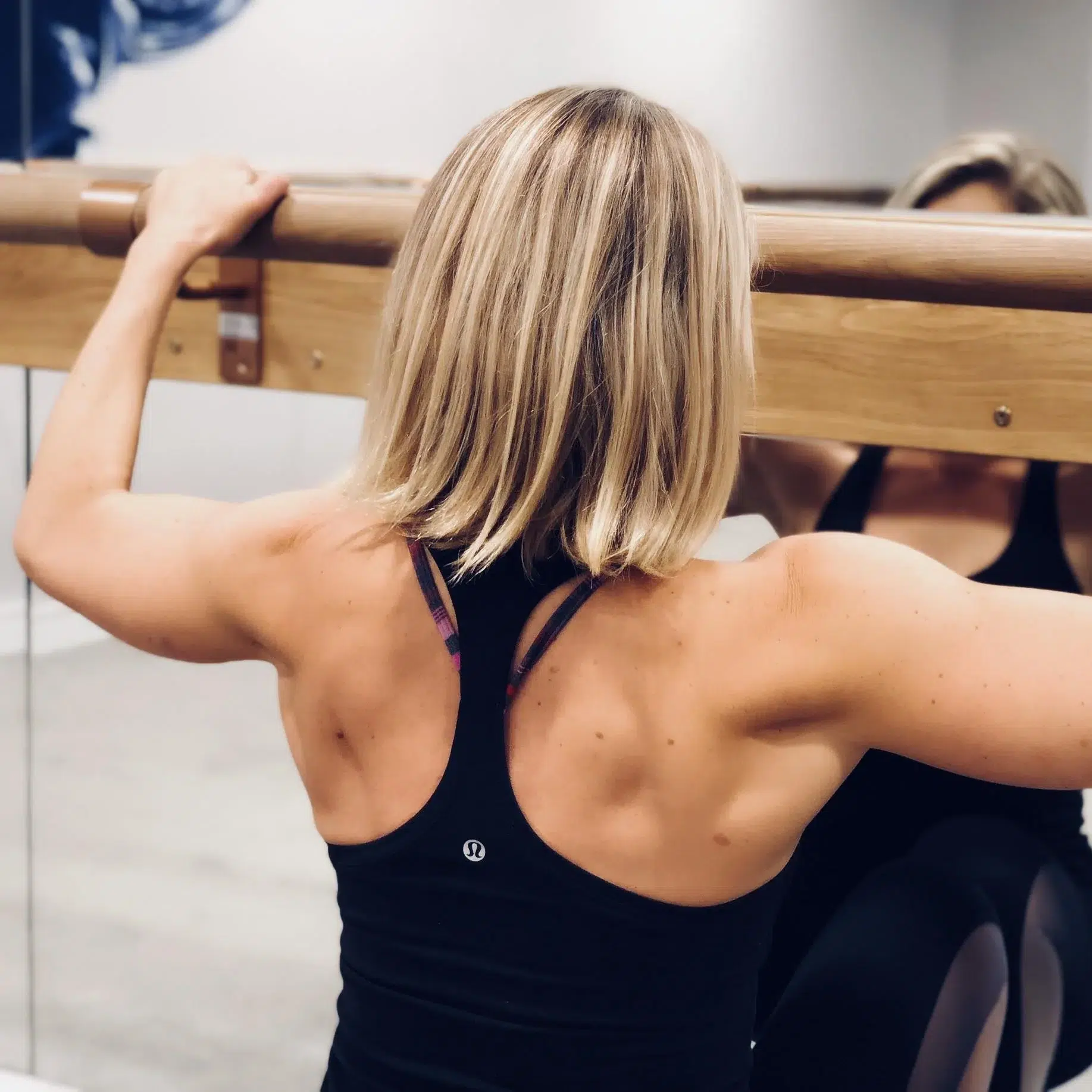 A female wearing a racer back sports top has her back to the camera displaying her back muscles whilst holding a ballet style bar in a barre class.