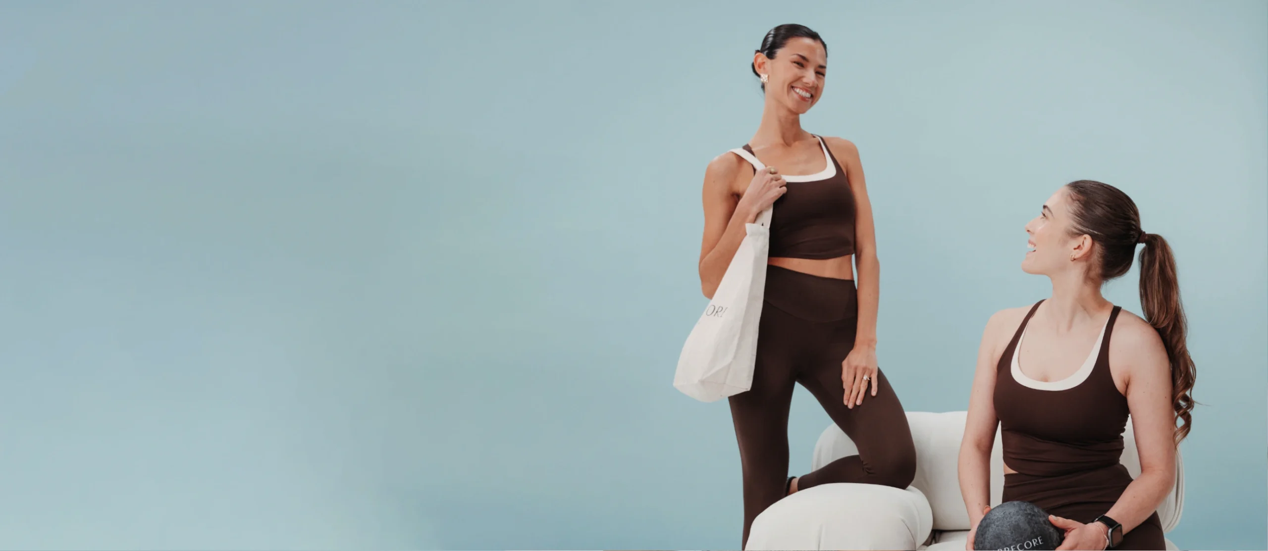 Two smiling women in matching brown Barrecore activewear, one carrying a Barrecore tote bag and the other holding a Barrecore ball