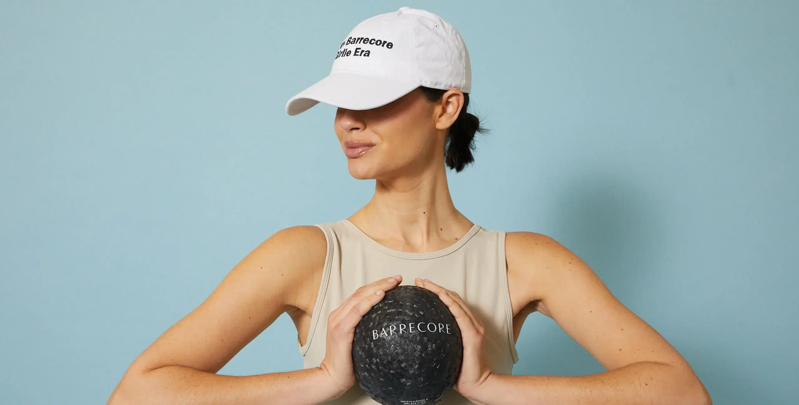 Woman in light activewear and white 'In My Barrecore Girlie Era' cap holding a branded Barrecore weighted ball against a pastel blue background
