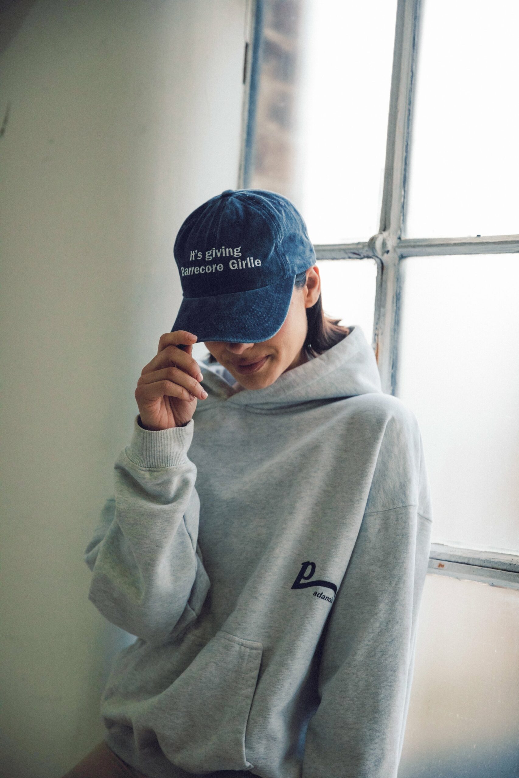 Woman in a hoodie wearing a navy cap embroidered with 'It’s giving Barrecore Girlie', showcasing branded fitness lifestyle merch.