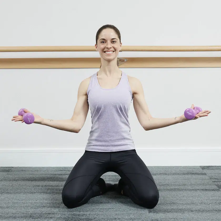 A female barre instructor sits on the floor of a barre studio with her knees bent and arms out wide to the sides holding two small weights