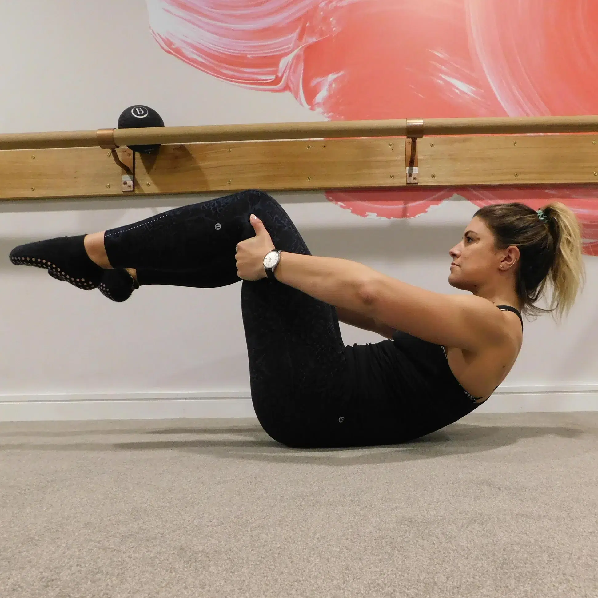 A female barre instructor is on the floor in a barre studio in a mid back curl position, her lower back is on the floor, her upper back, head and legs are raised.