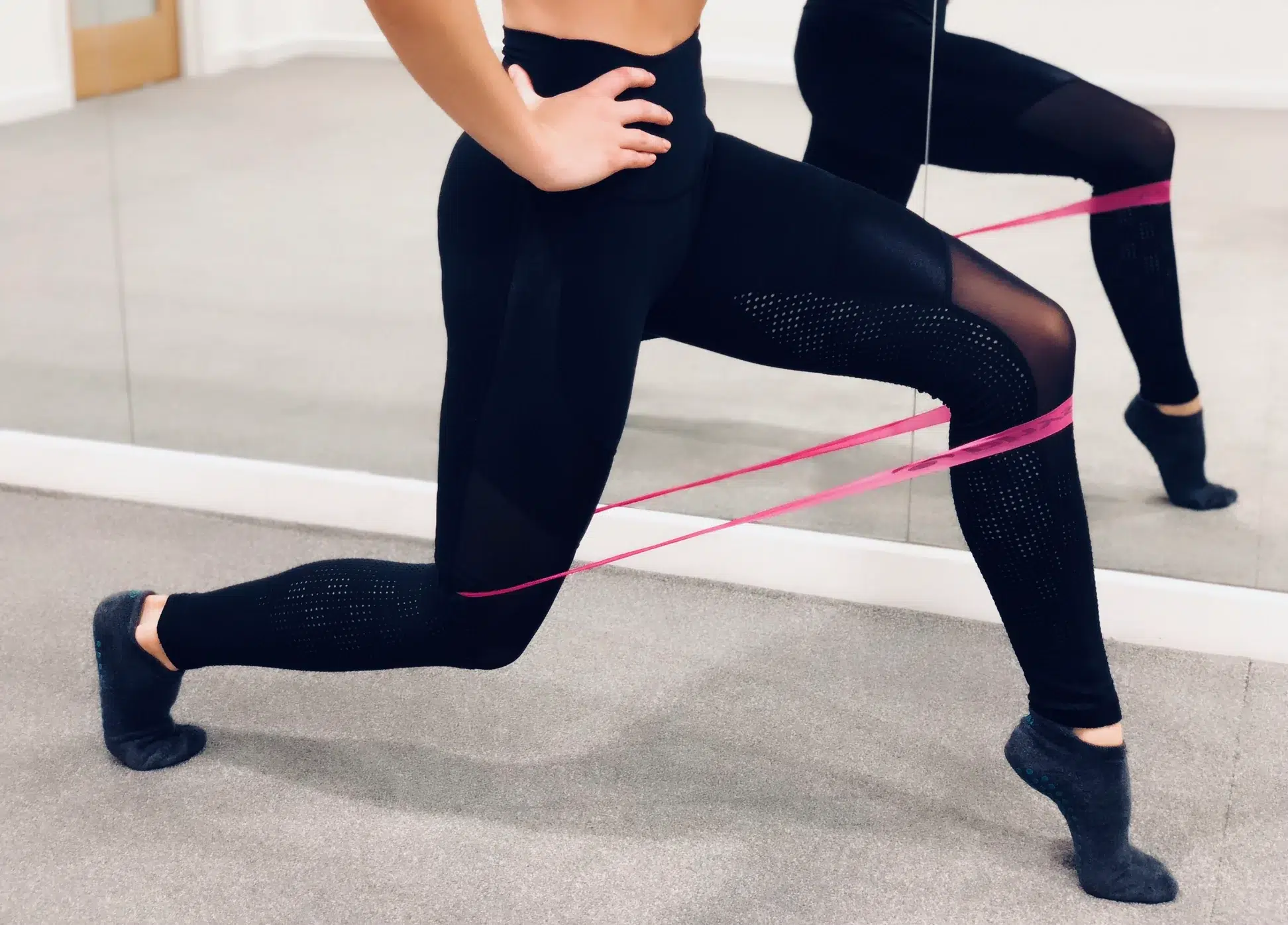 Female legs in a lunge position using a resistance band around both legs. There is a reflection in the wall mirror of a barre studio.