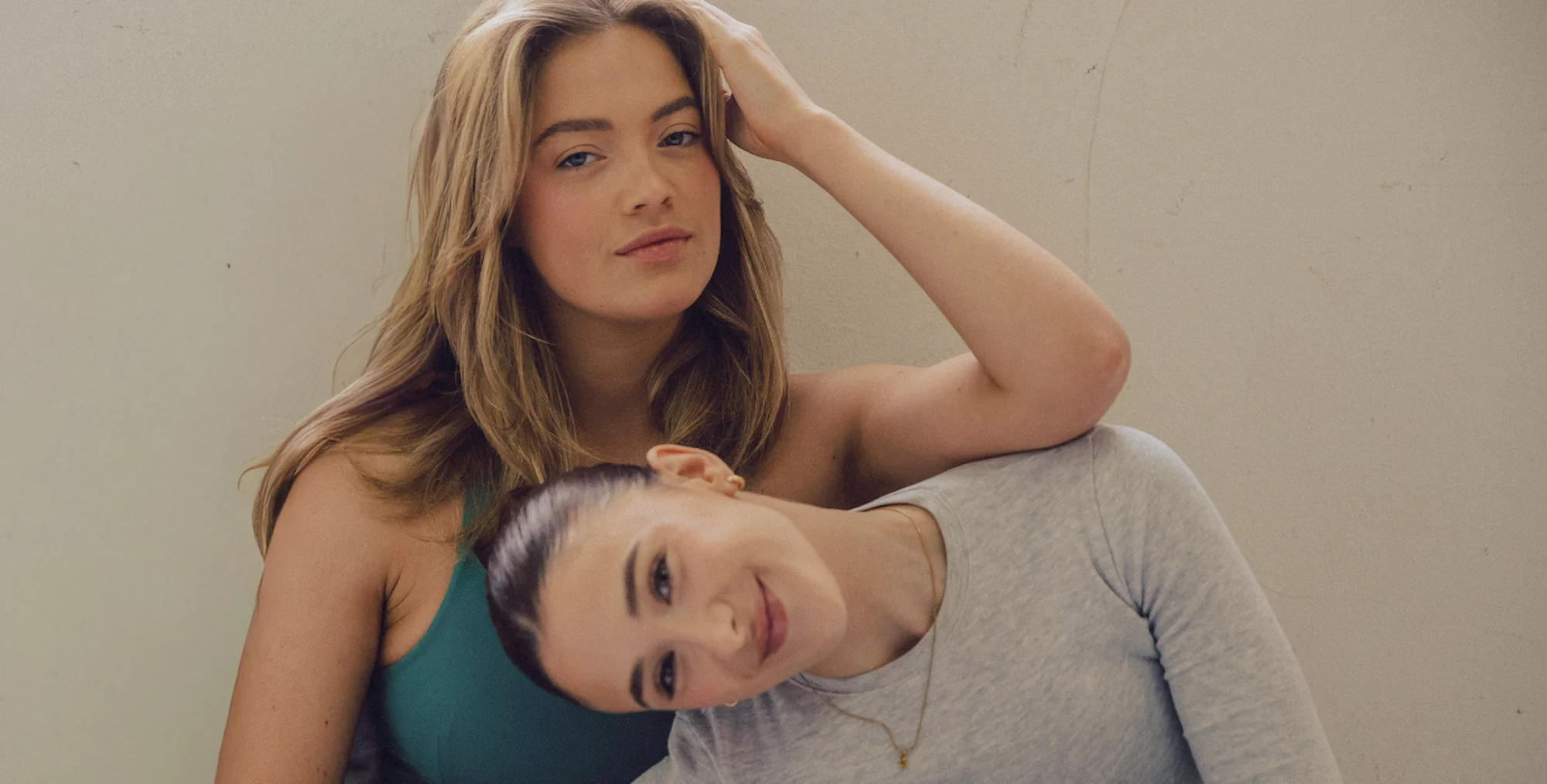 Two women in activewear posing together in soft natural light, one resting against the other's shoulder