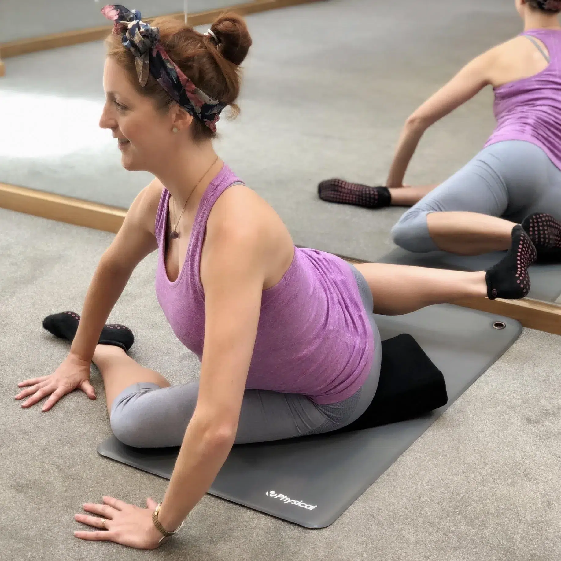 A female in a purple sports top and grey leggings is seated on the floor in a barre studio in a modified pretzel position with her hands supporting her.
