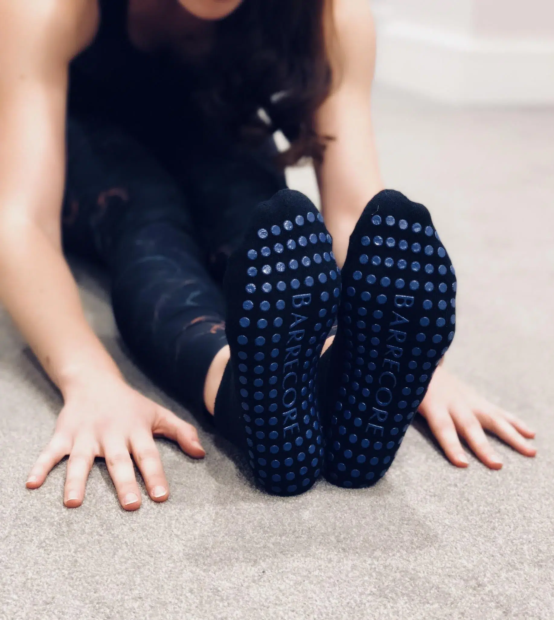 A female is sat on a carpeted floor with the soles of her feet showing, she is wearing black socks with blue grip dots and the word Barrecore