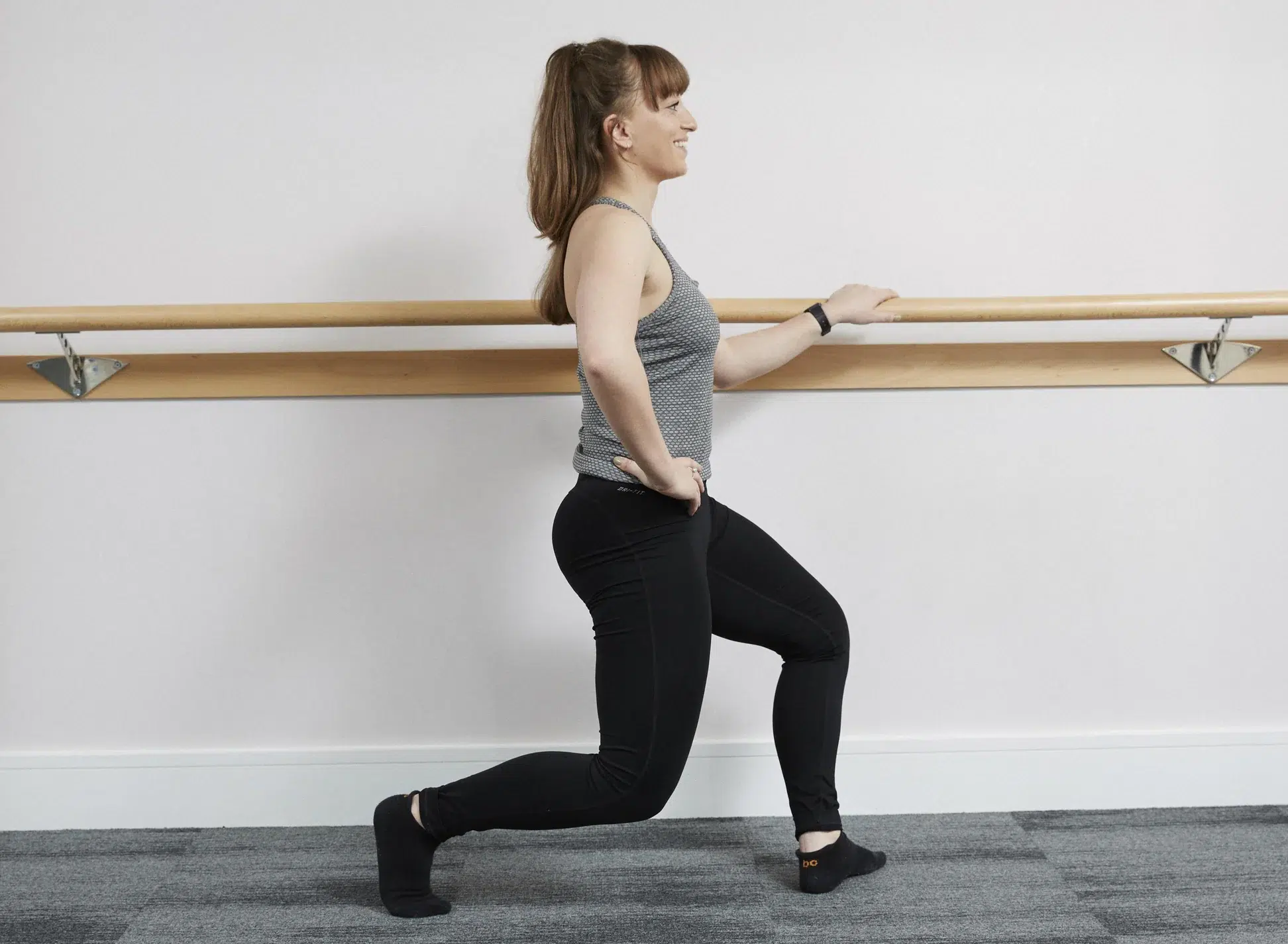 A female barre instructor is sideways on holding a barre and demonstrating a lunge position