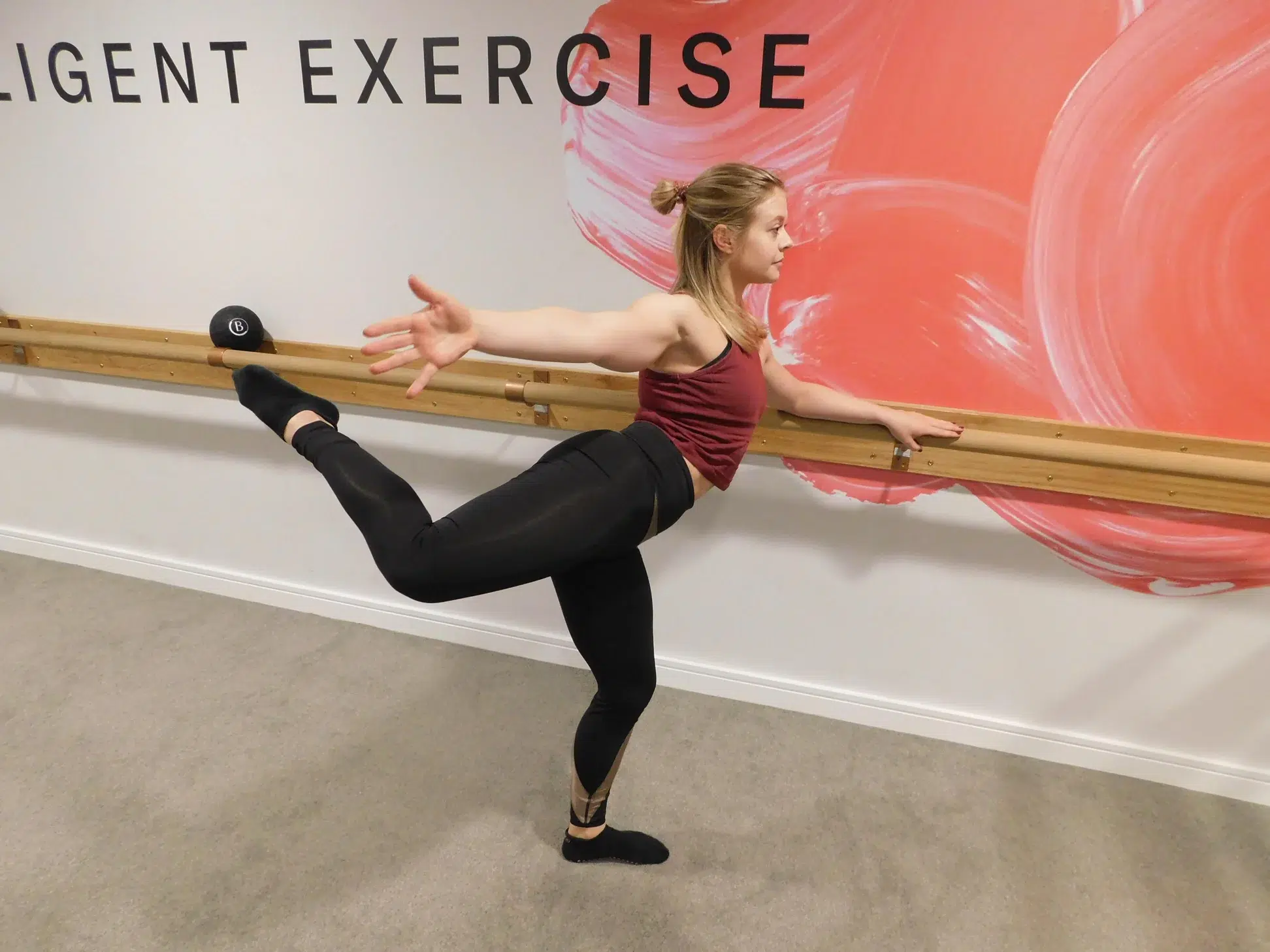 A female barre instructor is holding a ballet barre and holding her right leg up in a backward kick standing pretzel position