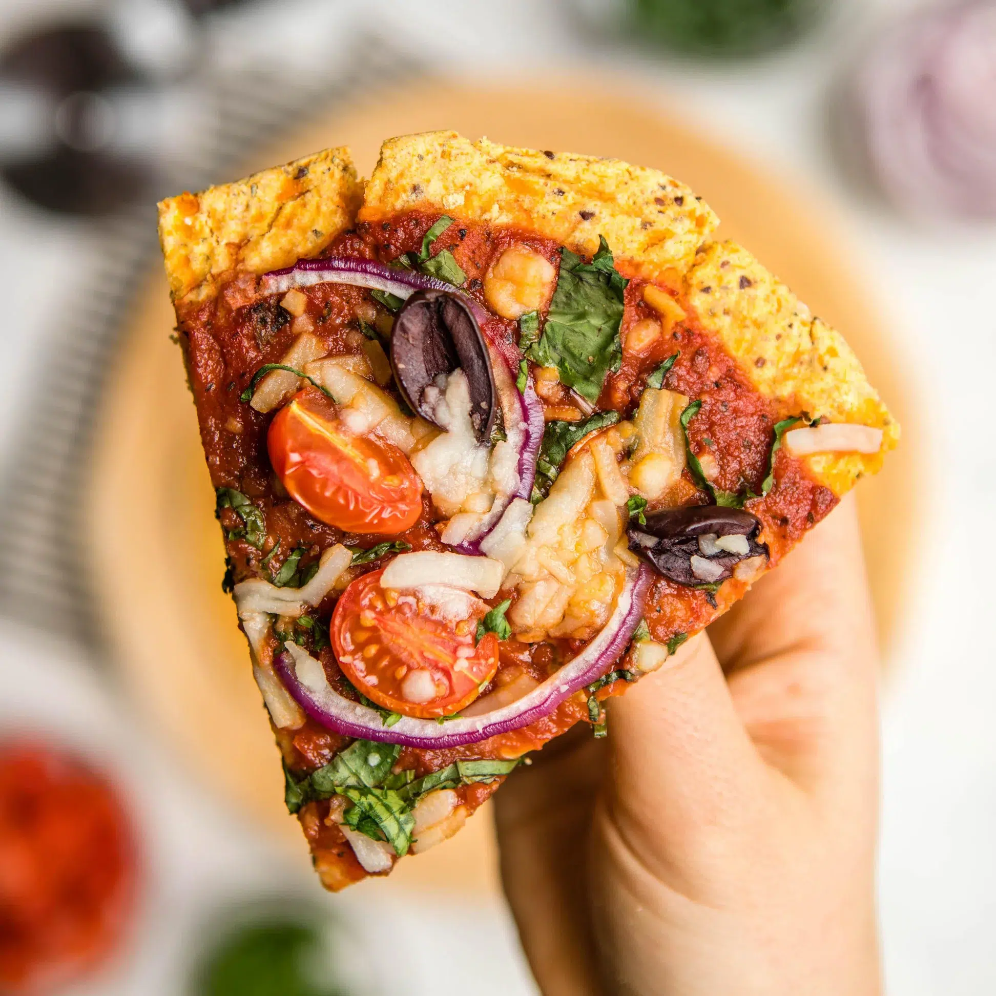 A slice of pizza in a hand with a sweet potato pizza base and a selection of toppings including tomatoes, cheese, olives and onion