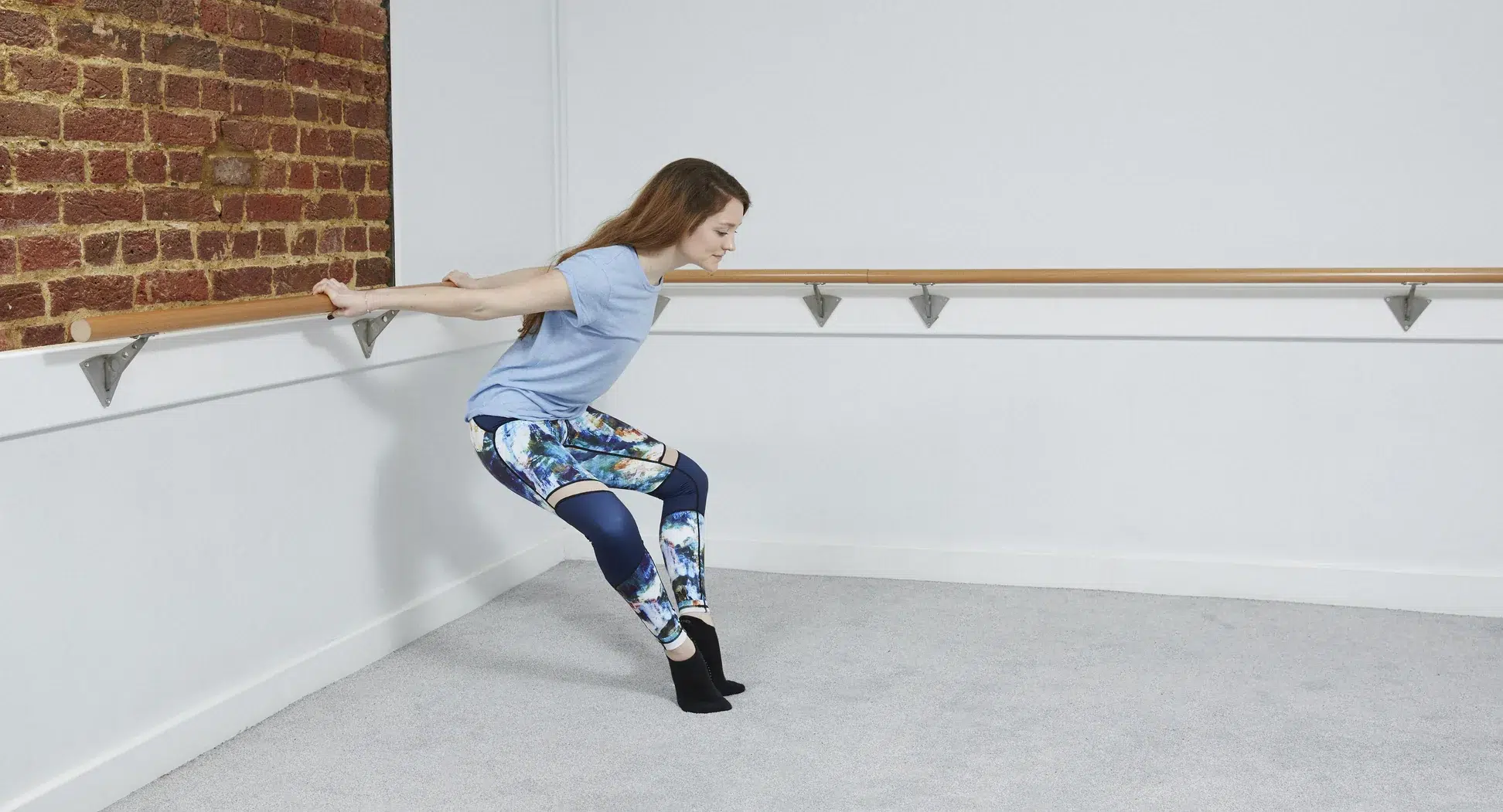A female barre instructor has her back to the barre and is holding it behind her to support a leg stretch