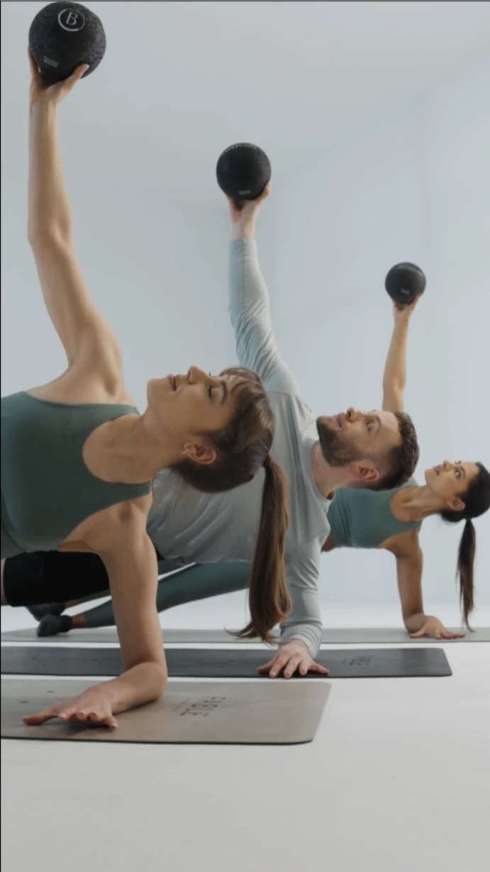 Barrecore clients in a signature class holding side planks with weighted balls, combining isometric strength, control, and precision in a refined, low-impact barre workout.