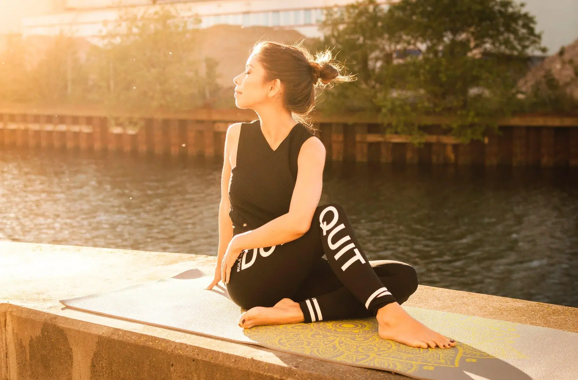 A female barre instructor is wearing sportswear and sat on the wall of a riverbank in a stretch position looking towards the sun.